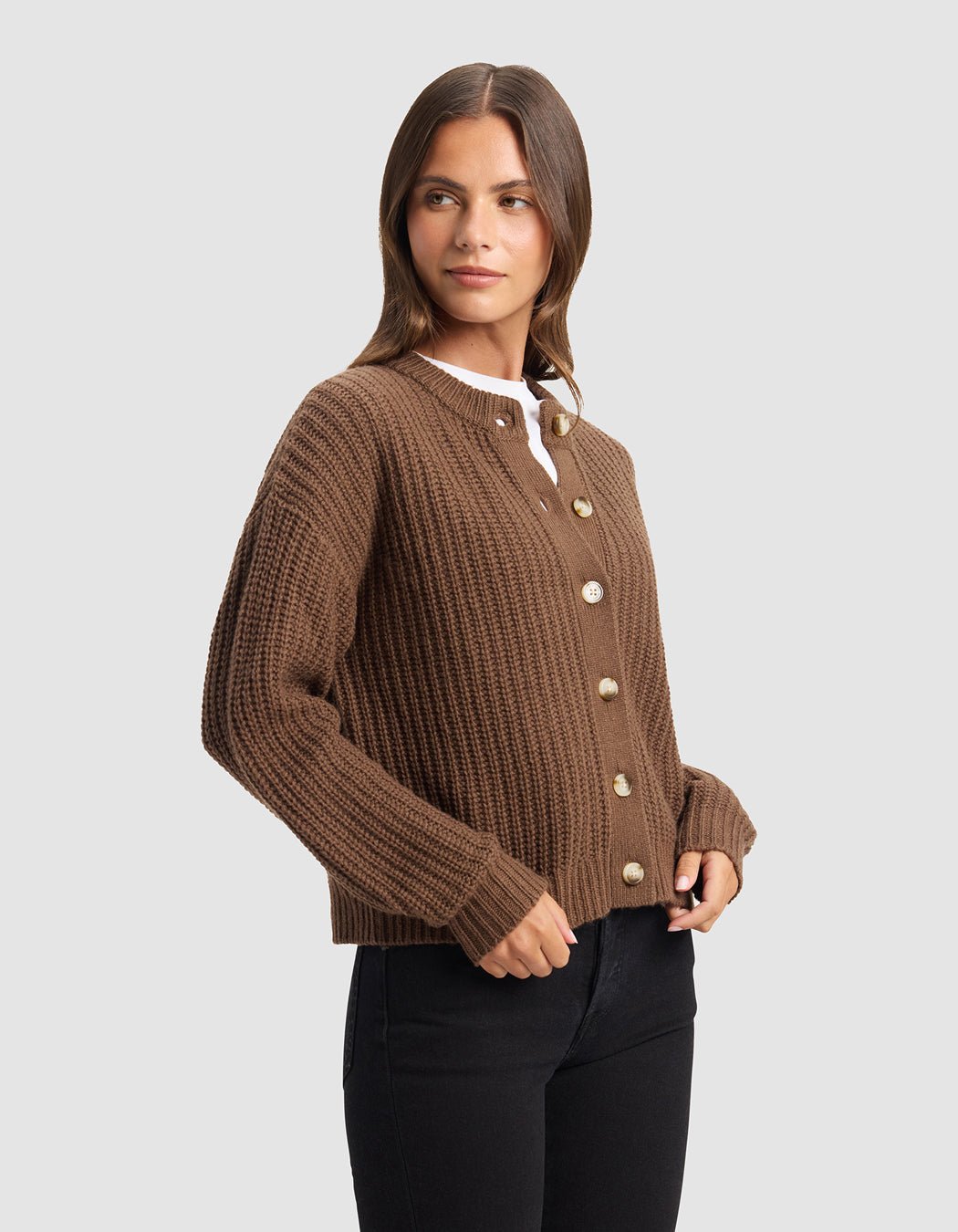 A woman with long brown hair models the Cozy Earth Women's Arden Cardigan, a brown ribbed button-up, layered over a white top and black pants against a plain light gray background.|Color:Cedar