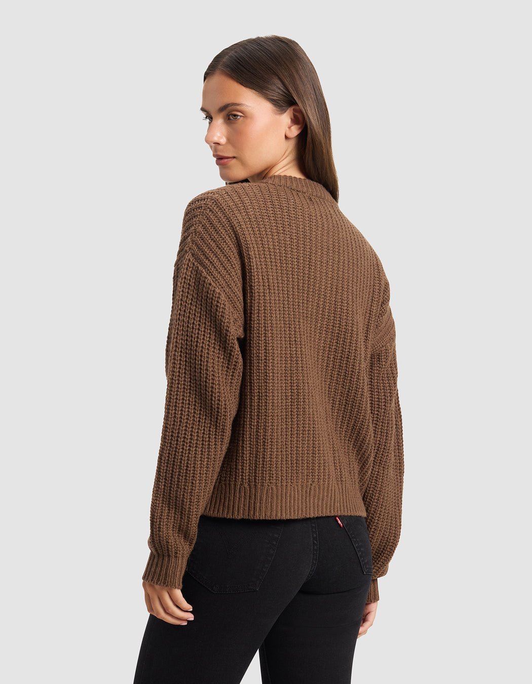 A woman with long brown hair models the Cozy Earth Women's Arden Cardigan, standing against a plain light gray background and looking over her shoulder toward the camera.