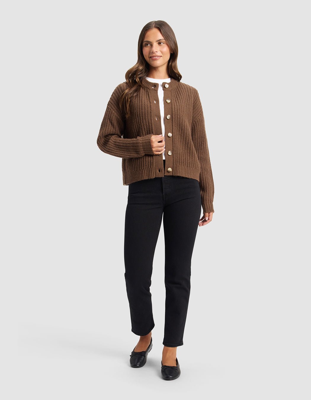 A woman with long brown hair stands against a light gray background, wearing the Cozy Earth Women's Arden Cardigan in brown over a white top, paired with black pants and flats. She smiles softly while gazing to the side.