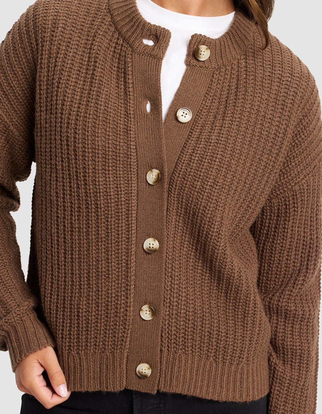 A person wears Cozy Earth's Women's Arden Cardigan, a brown ribbed knit with large tan buttons, partially buttoned over a white shirt and black pants. Their left hand rests near the bottom button.