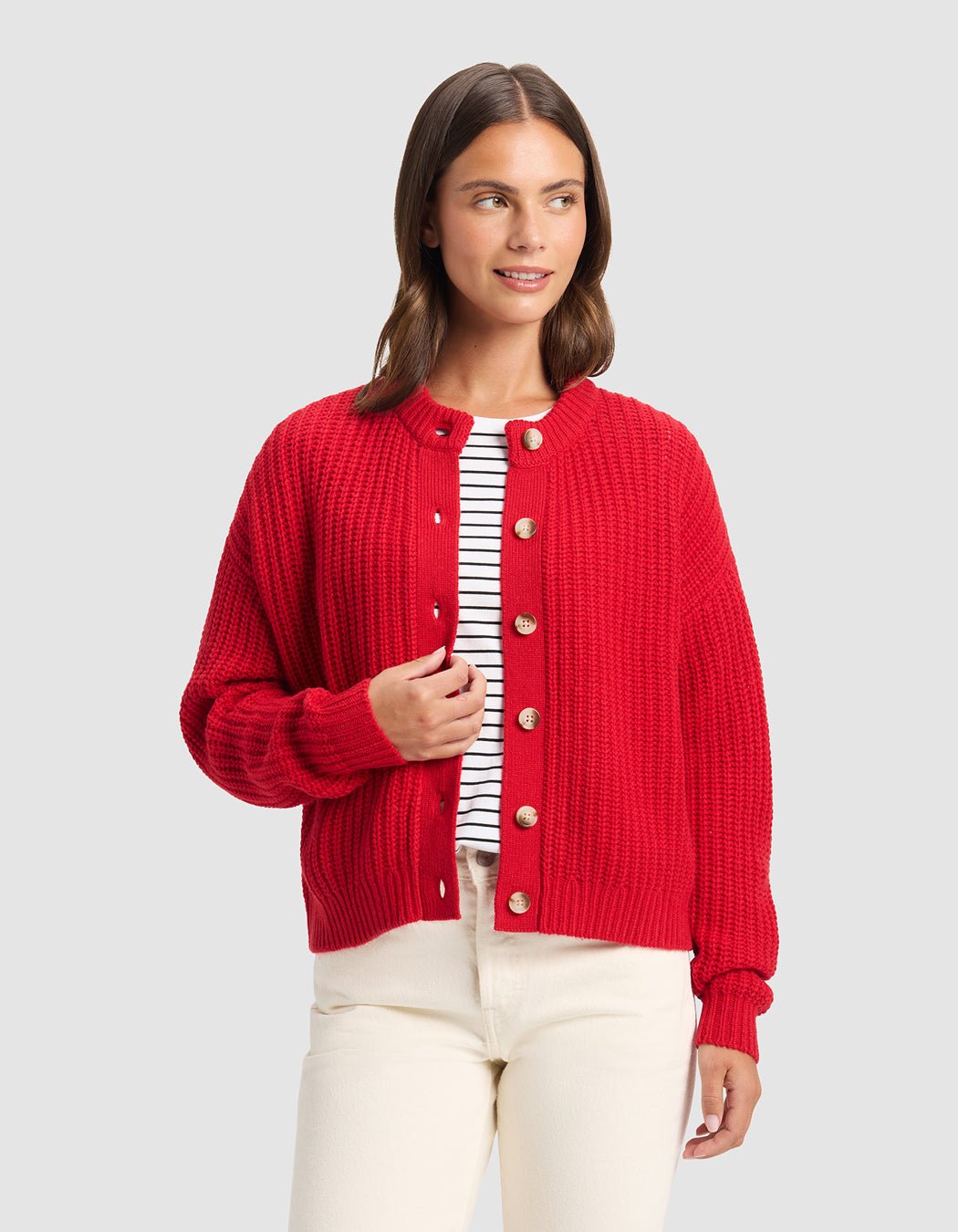 A woman wears the Cozy Earth Women's Arden Cardigan in bright red over a black and white striped shirt with cream pants, standing against a light gray background and smiling slightly while looking to her left.