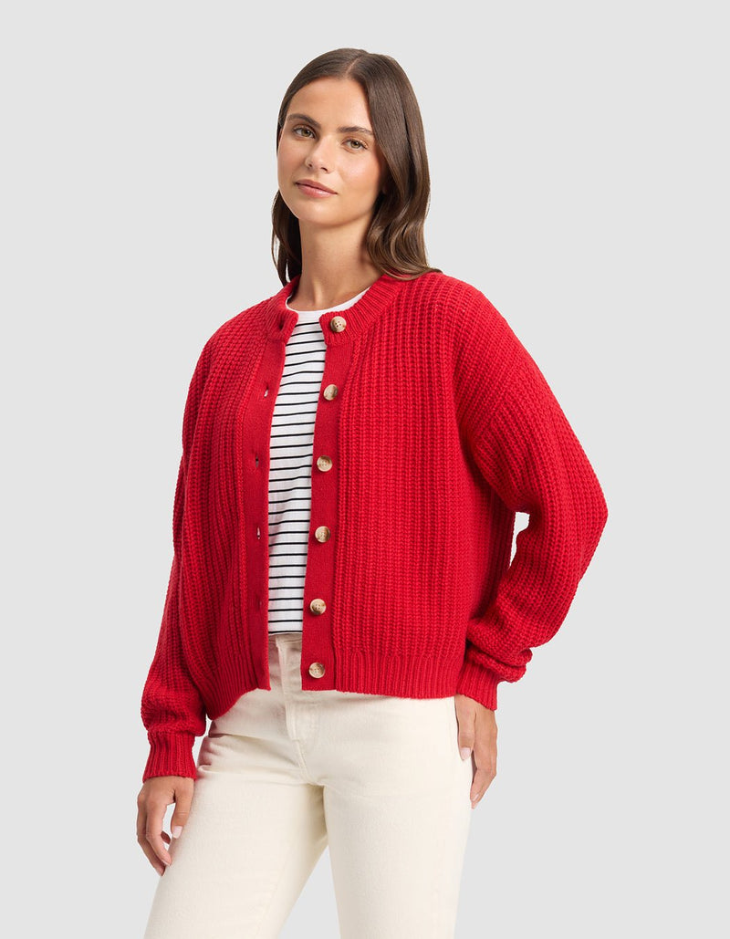 A woman with straight brown hair and a relaxed expression wears the Cozy Earth Women's Arden Cardigan in red over a white shirt with black stripes and cream-colored pants, standing against a plain background.