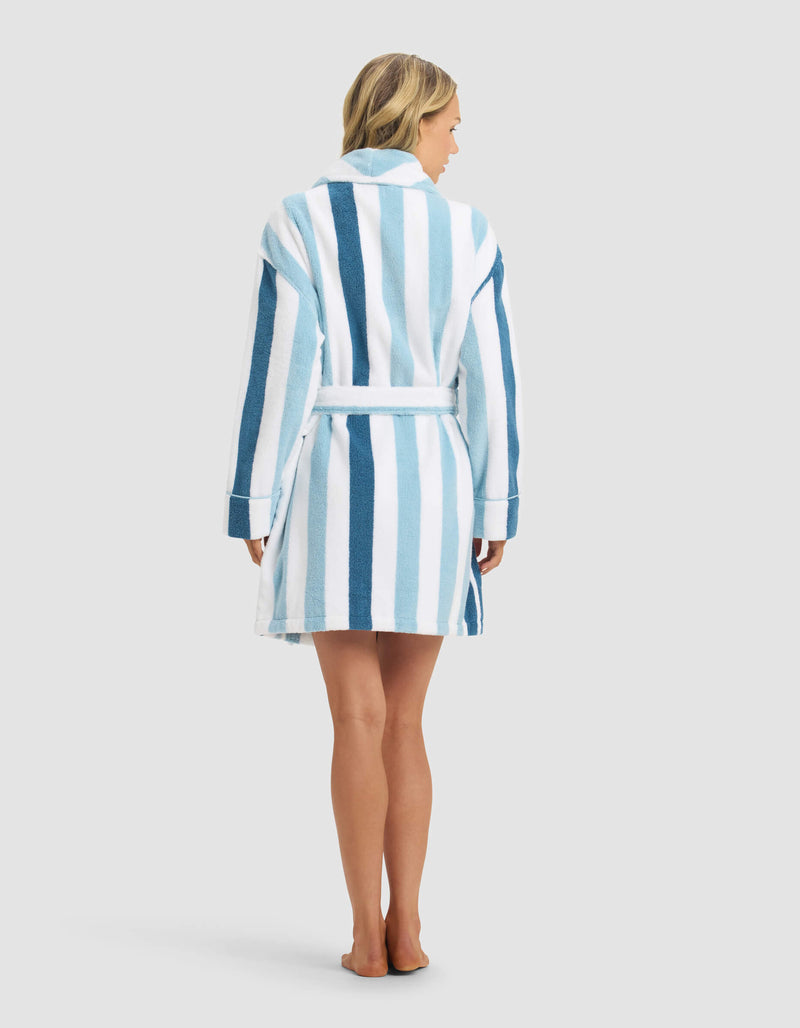 A woman with loose blonde hair stands barefoot, facing away, wearing the Cozy Earth Classic Stripe Resort Robe—knee-length with vertical blue and white stripes, a belted waist, against a plain light gray background.