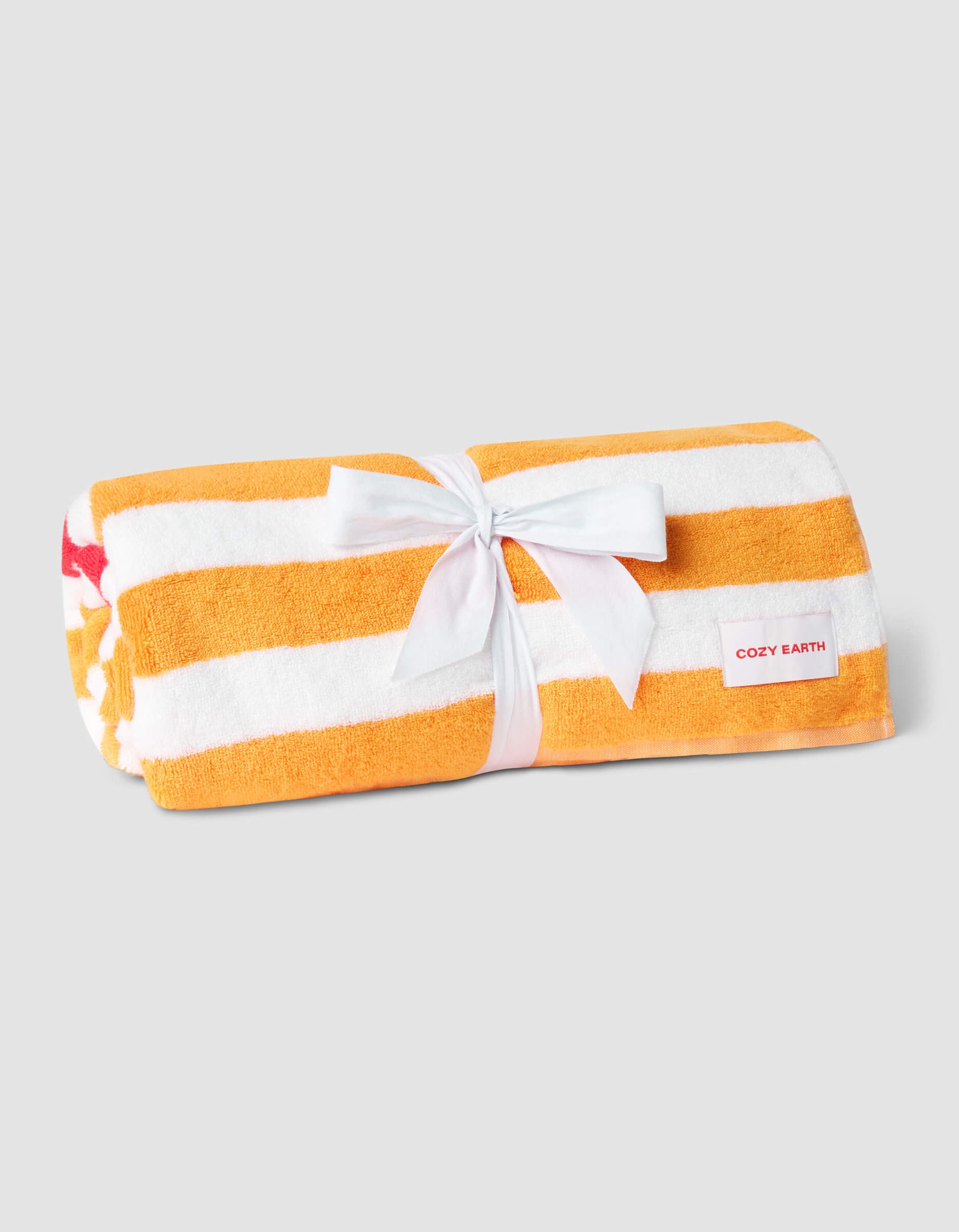 The Cozy Earth Classic Stripe Resort Towel features wide orange and white stripes, tied with a white ribbon. A red "Cozy Earth" label accents one end, all set against a plain light gray background.