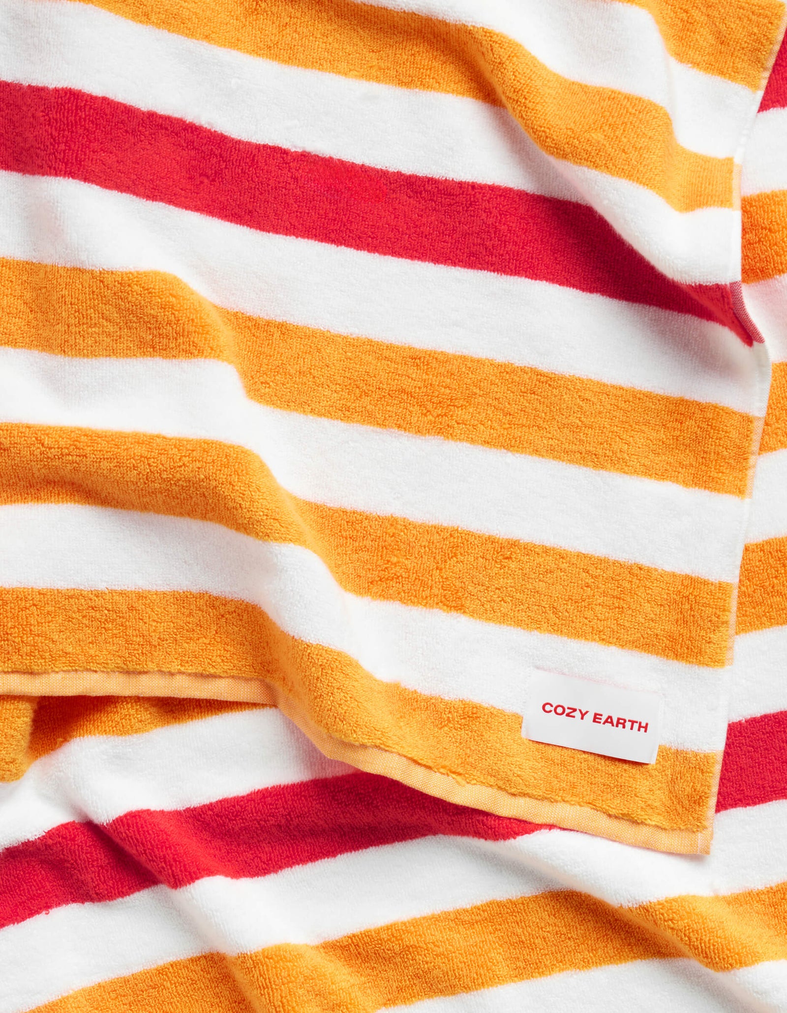 The Classic Stripe Resort Towel by Cozy Earth features bold orange, red, and white horizontal stripes and a soft feel. A small corner tag displays the Cozy Earth brand in red text.