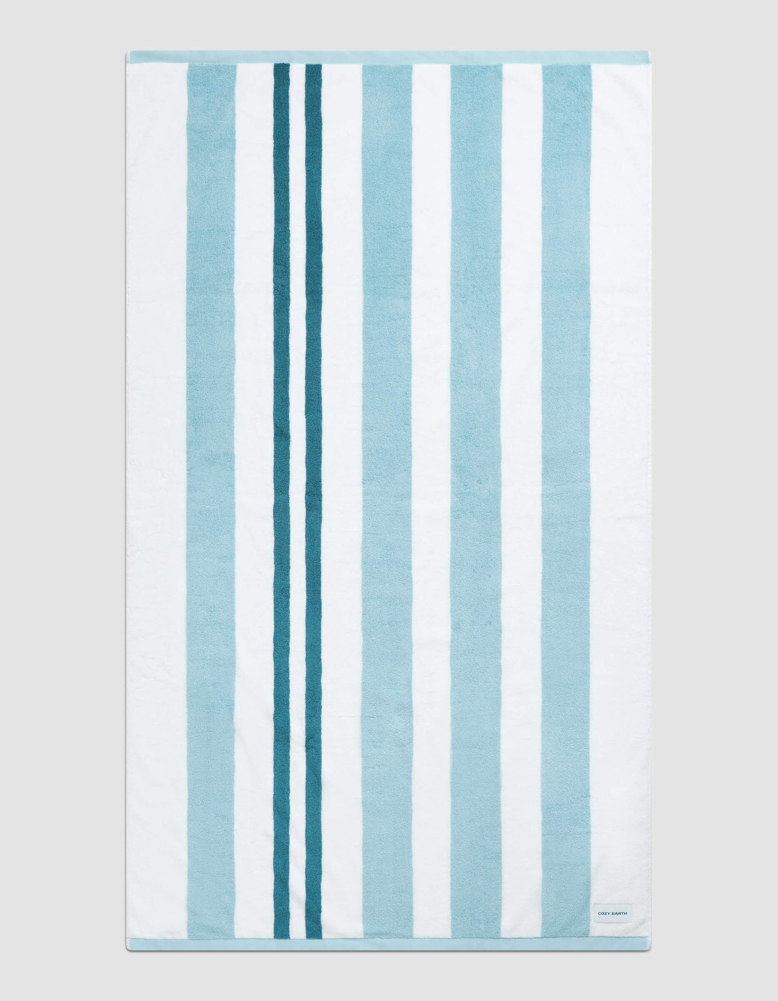 The Classic Stripe Resort Towel by Cozy Earth in Wave Stripe is shown laid flat on a light gray background. Alternating vertical blue and white stripes and a small white label in the bottom right corner are visible.