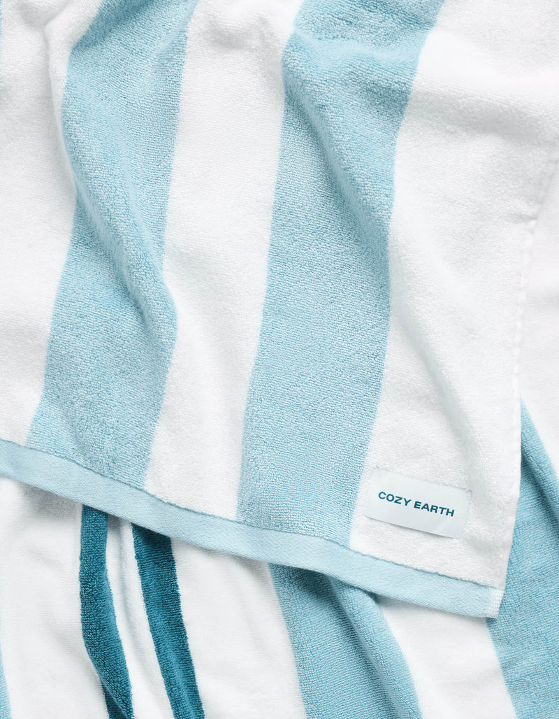 A close-up of the Cozy Earth Classic Stripe Resort Towel, showcasing its soft texture and wide white and light blue vertical stripes, with a small "COZY EARTH" label in the corner.