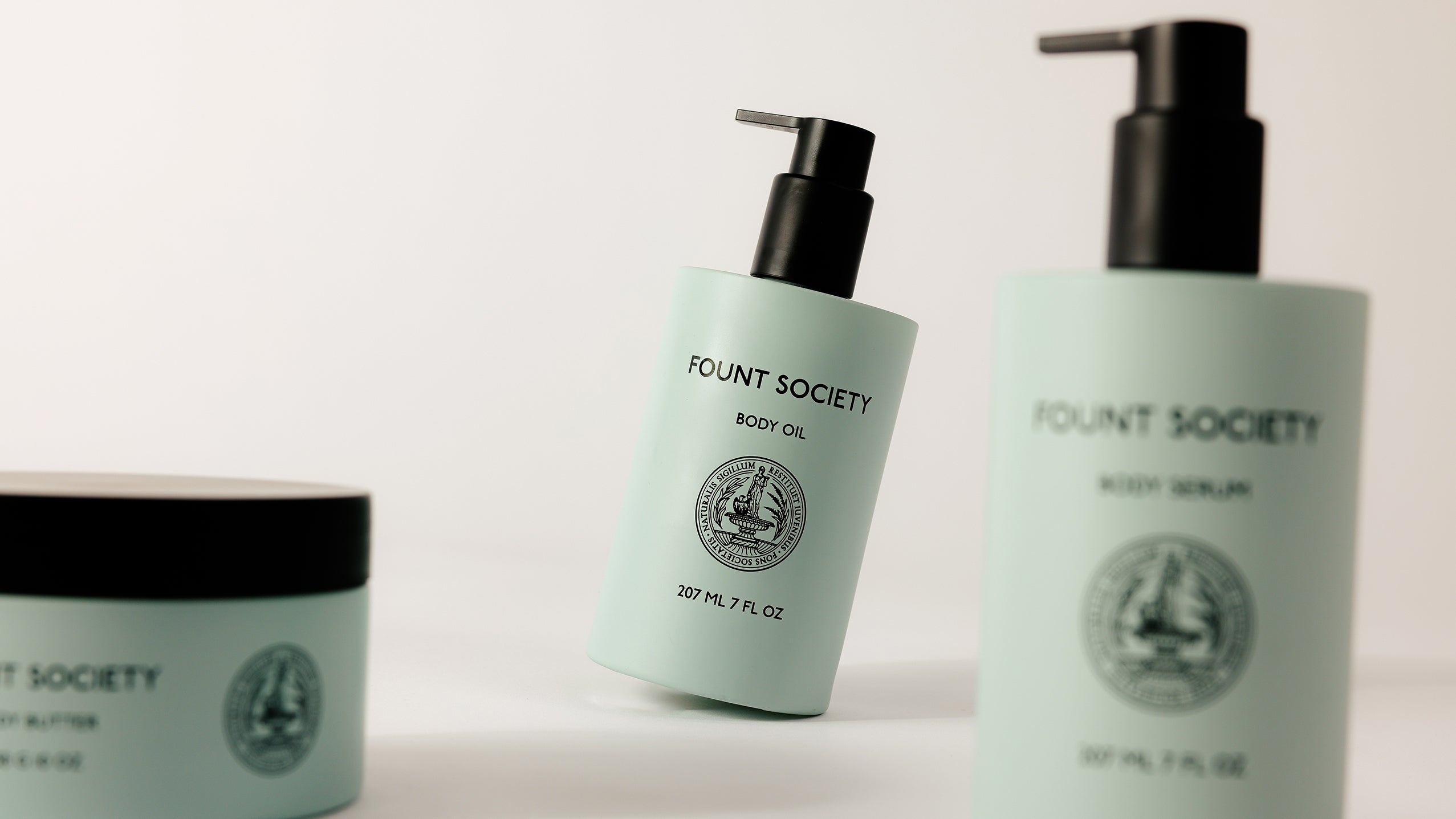 Image of three Cozy Earth Moisture Trio products with light blue packaging. The focus is on a body oil bottle with a pump dispenser in the center, slightly tilted. Two other products, a body serum and a jar, are partially visible on the sides.