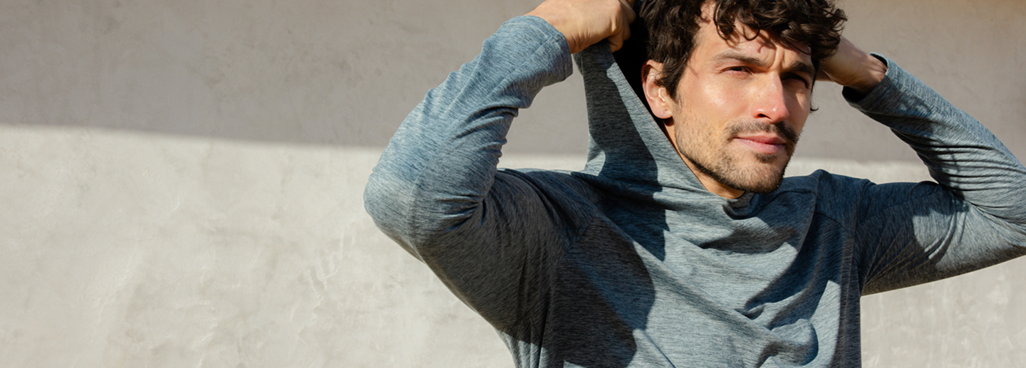 A person stands outdoors, wearing a gray hoodie and pulling the hood over their head. They are looking into the distance with a thoughtful expression. The background is a plain, sunlit wall.
