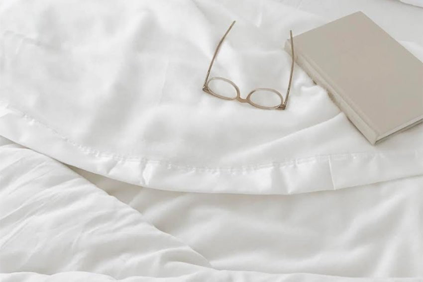 A neatly made white bed with a pair of glasses and a beige book placed on top of the Cozy Earth Bamboo Blanket. The scene suggests a calm, peaceful ambiance, possibly right before or after reading in bed. 