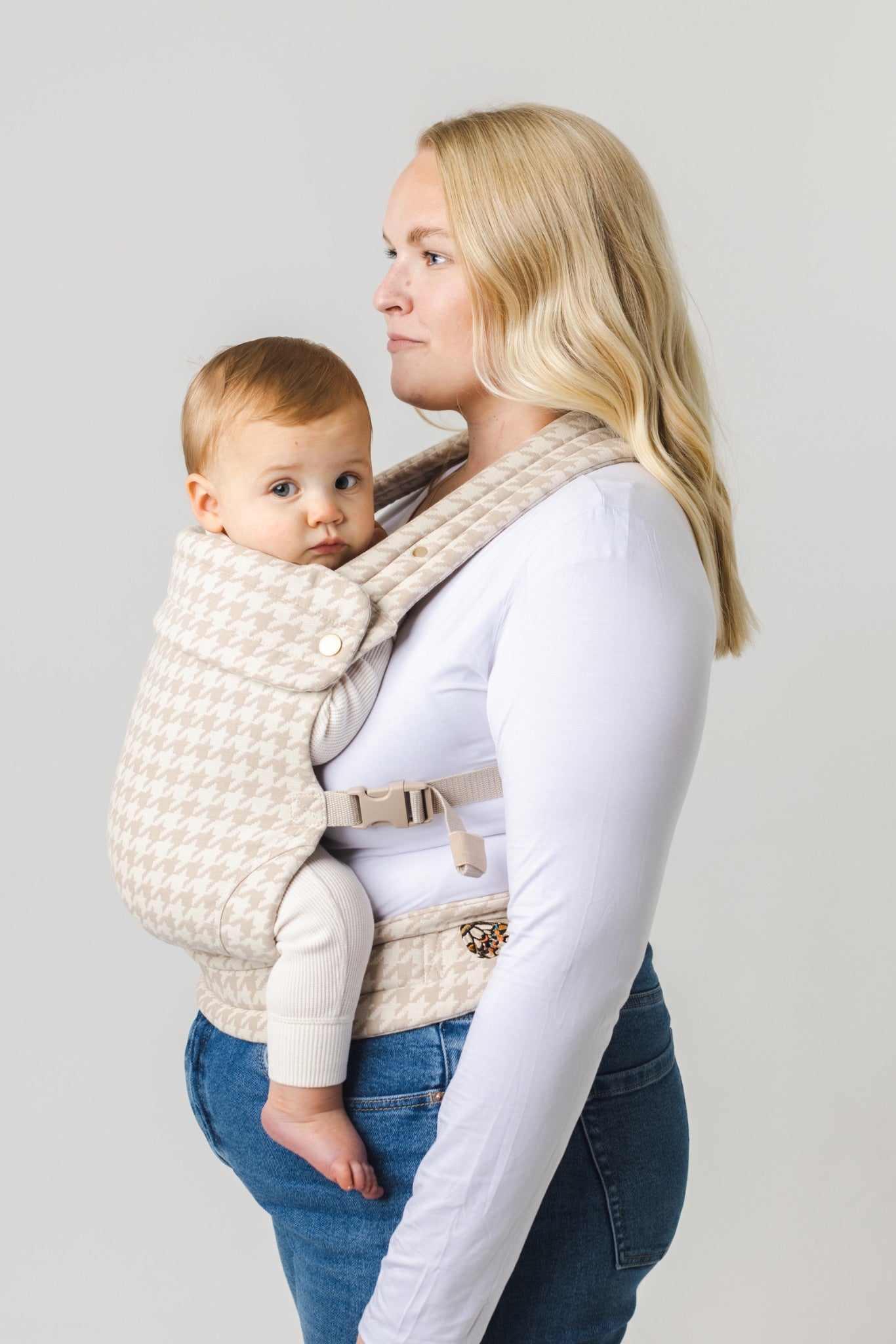 A woman with long blonde hair wears a white shirt and blue jeans while carrying a baby in the Mabē The Monarch Carrier - Houndstooth. Both face right against a plain background.