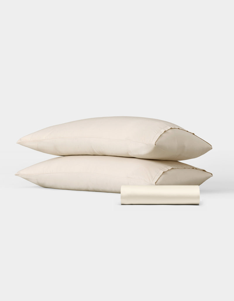 The Bamboo Bedding Basic Bundle by Cozy Earth features two cream-colored pillows stacked together with a matching rolled pillowcase in front, displayed against a plain white background.