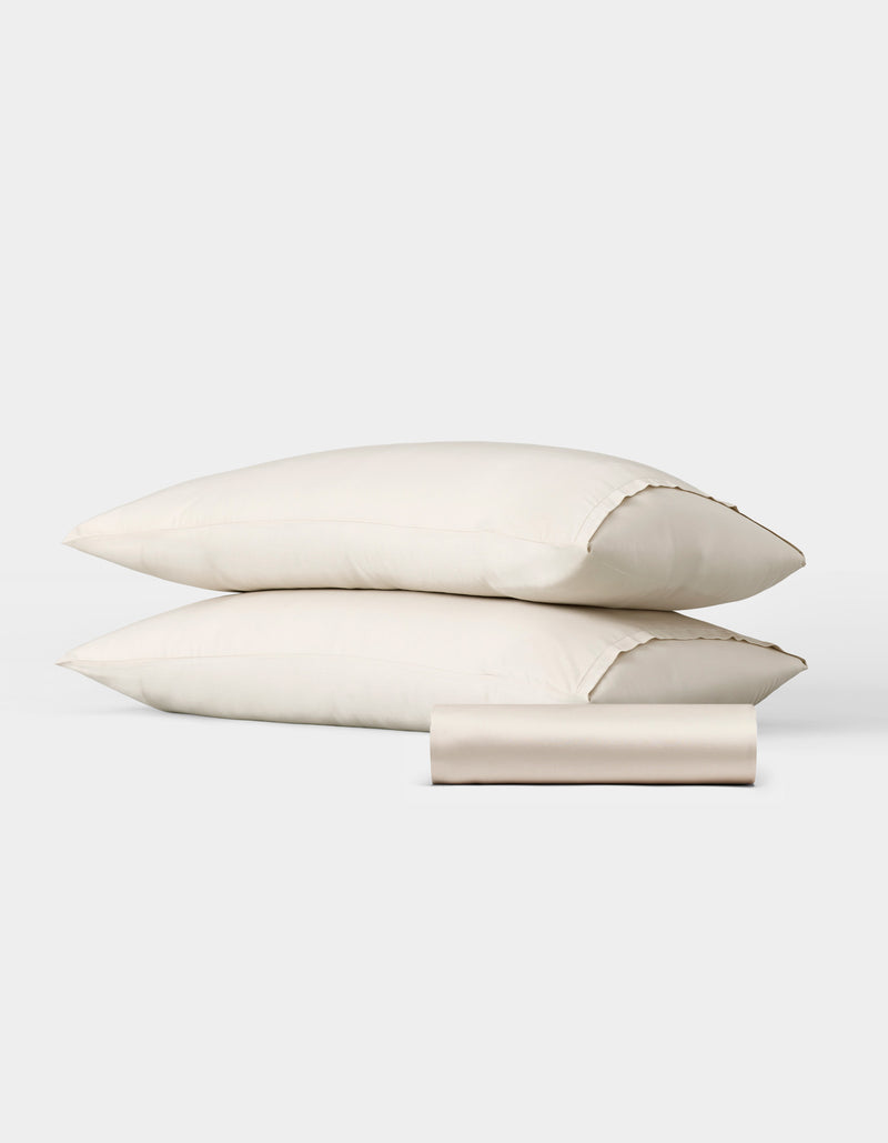 The Bamboo Bedding Basic Bundle by Cozy Earth features two cream-colored pillows stacked horizontally with a matching rolled-up fabric case in front, all displayed against a plain white background.