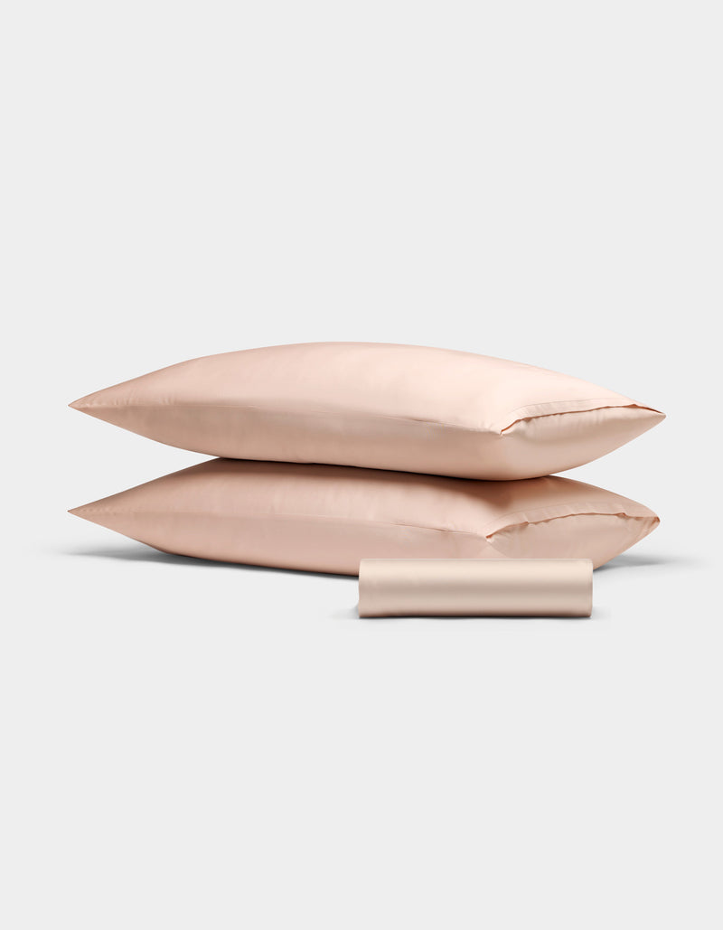 The Cozy Earth Bamboo Bedding Basic Bundle features two light pink satin pillowcases stacked, with a matching satin sleep mask in front, all set against a plain white background.
