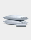 The Bamboo Bedding Basic Bundle by Cozy Earth features two light gray pillowcases stacked together and a matching rolled-up fabric piece in front, all displayed against a plain light gray background.|Color:Shore