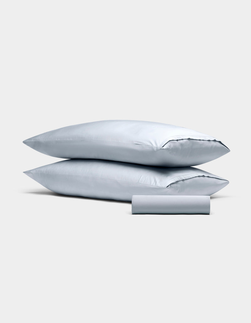 The Bamboo Bedding Basic Bundle by Cozy Earth features two light gray pillowcases stacked together and a matching rolled-up fabric piece in front, all displayed against a plain light gray background.