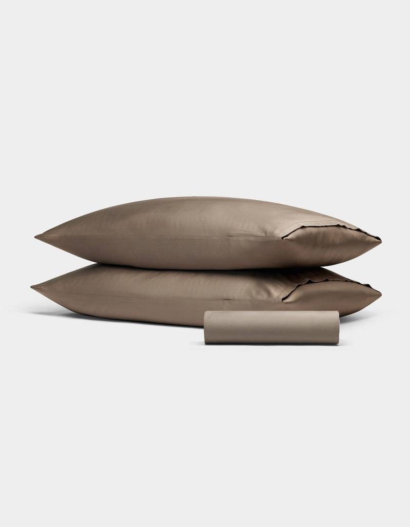 Two taupe Bamboo Bedding Basic Bundle pillowcases by Cozy Earth are stacked together, with a matching cylindrical fabric pouch in front, all set against a plain light gray background.