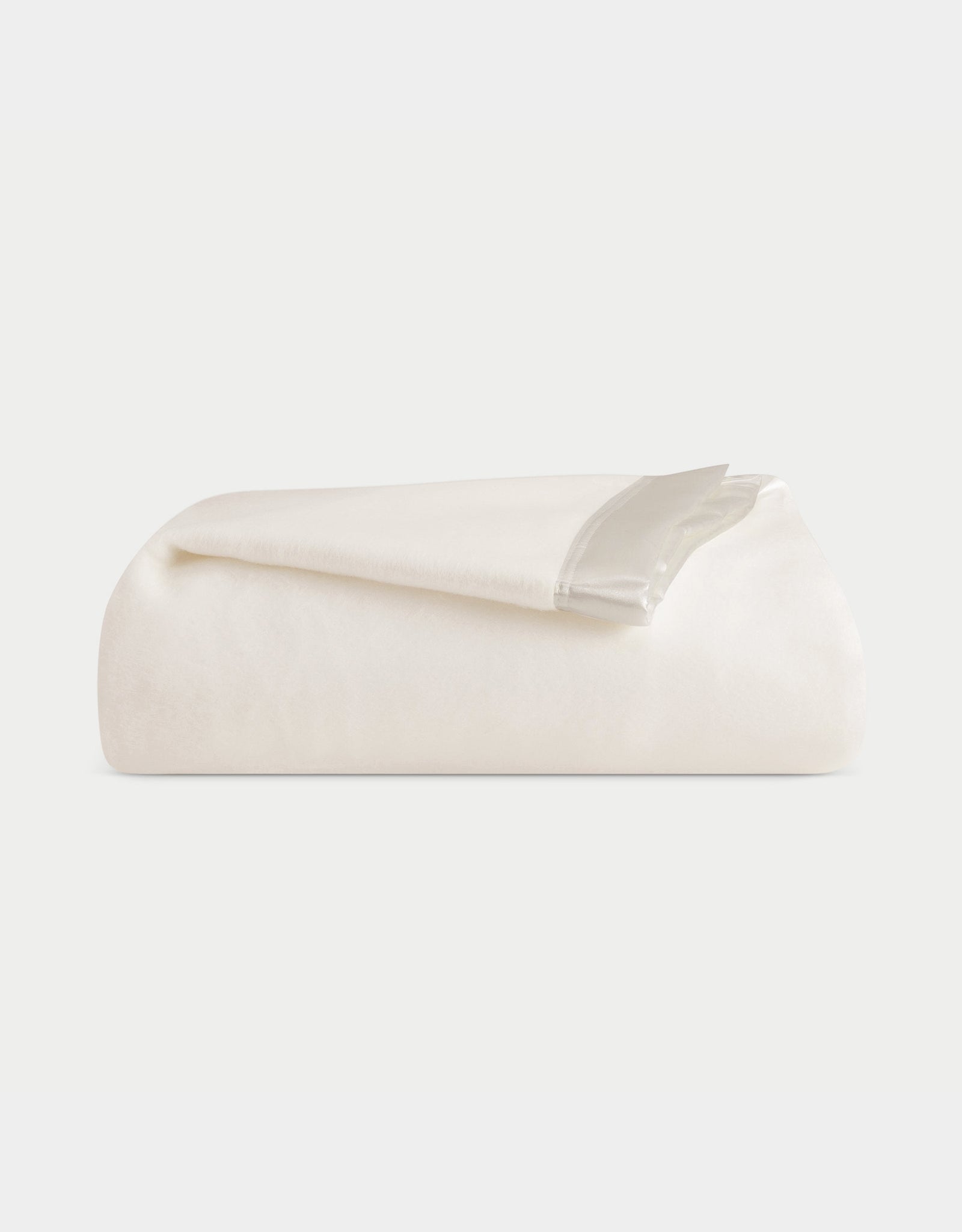 A neatly folded Cozy Earth Bamboo Blanket, soft in ivory, rests on a white surface. It features an elegant satin trim along one edge, enhancing its simple design. 