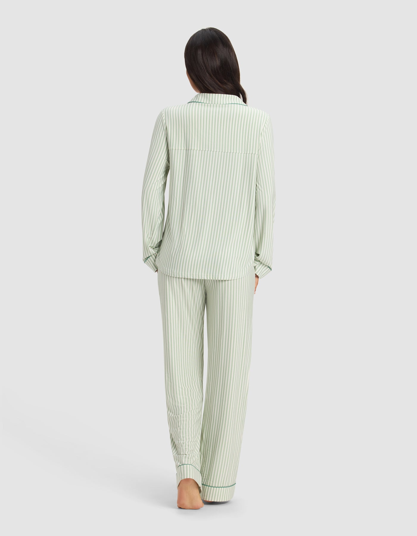 A person with long dark hair, barefoot, stands with their back to the camera, wearing the Cozy Earth x Divi: The Limited-Edition Dream Set—light green and white striped long-sleeve pajamas. The background is plain and light-colored.