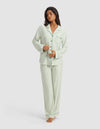 A woman stands barefoot against a white background, buttoning the limited-edition Cozy Earth x Divi Dream Set—light green and white striped long-sleeve pajamas and pants from Cozy Earth.