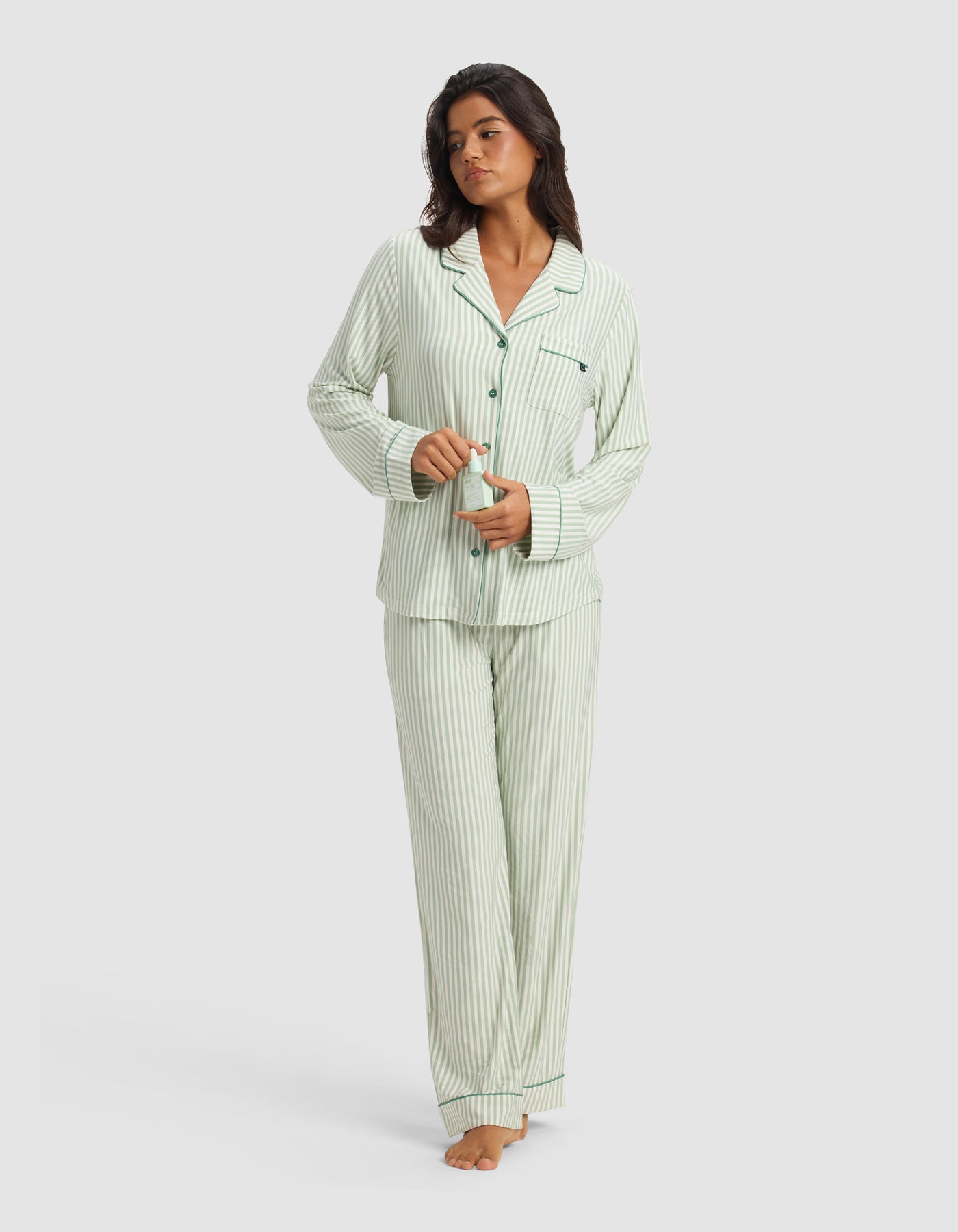 A woman stands barefoot against a white background, buttoning the limited-edition Cozy Earth x Divi Dream Set—light green and white striped long-sleeve pajamas and pants from Cozy Earth.