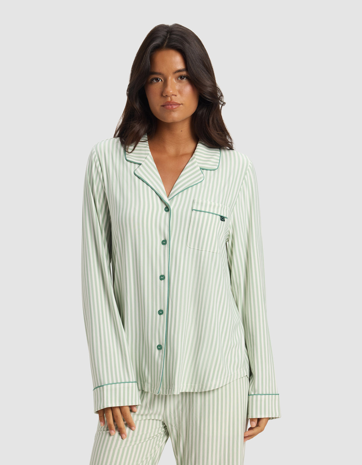 A woman models the Cozy Earth x Divi: The Limited-Edition Dream Set by Cozy Earth—light green and white striped pajamas with dark green buttons and piping—standing against a plain background with a neutral expression.