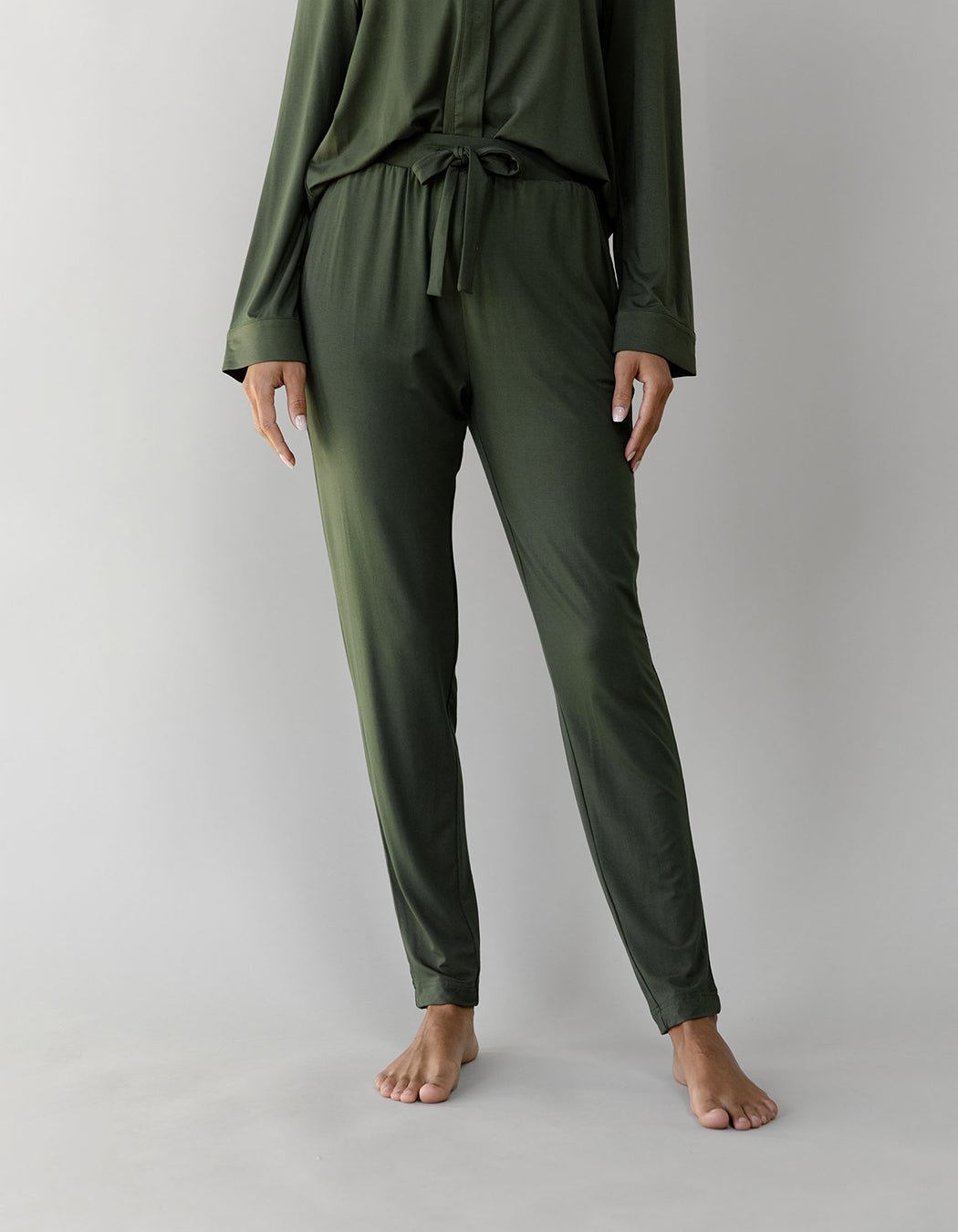 A person stands barefoot against a neutral backdrop, wearing the Cozy Earth Women's Bamboo Stretch-Knit Tapered Pant in green. The outfit includes a long-sleeve top and tie waist. Their face is not shown, keeping them unidentifiable. |Color:Olive