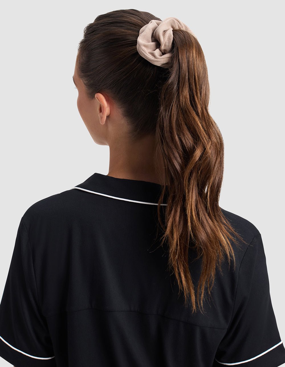 A woman with long, wavy brown hair in a ponytail, secured with a Cozy Earth Bamboo Rib-Knit Scrunchie in beige, is seen from behind. She wears a black shirt with white piping on the collar and sleeves against a light gray background.