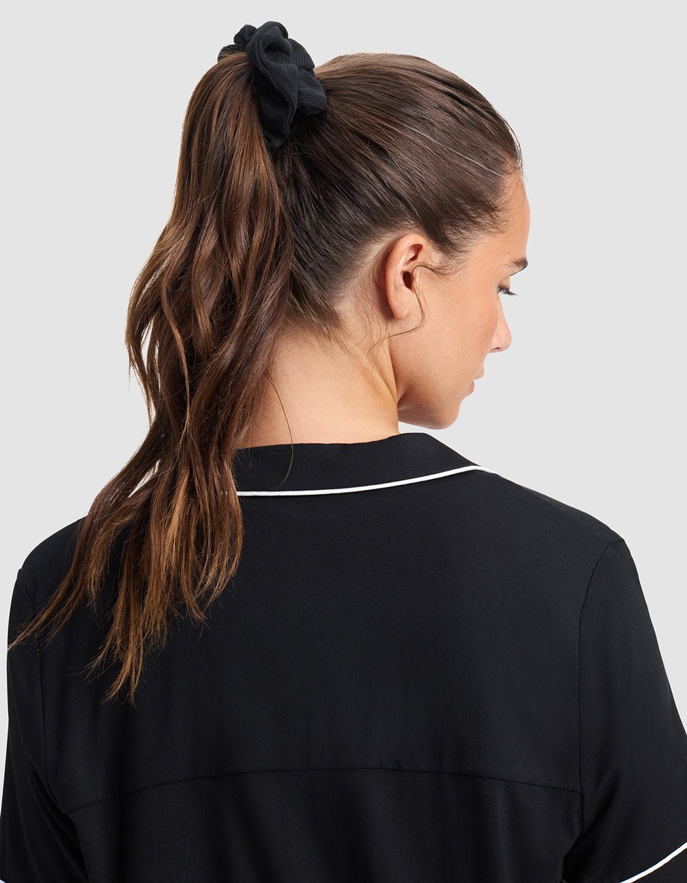 A woman with wavy brown hair in a high ponytail, secured by a Cozy Earth Bamboo Rib-Knit Scrunchie, wears a black top with white piping and is shown from behind against a light gray background.