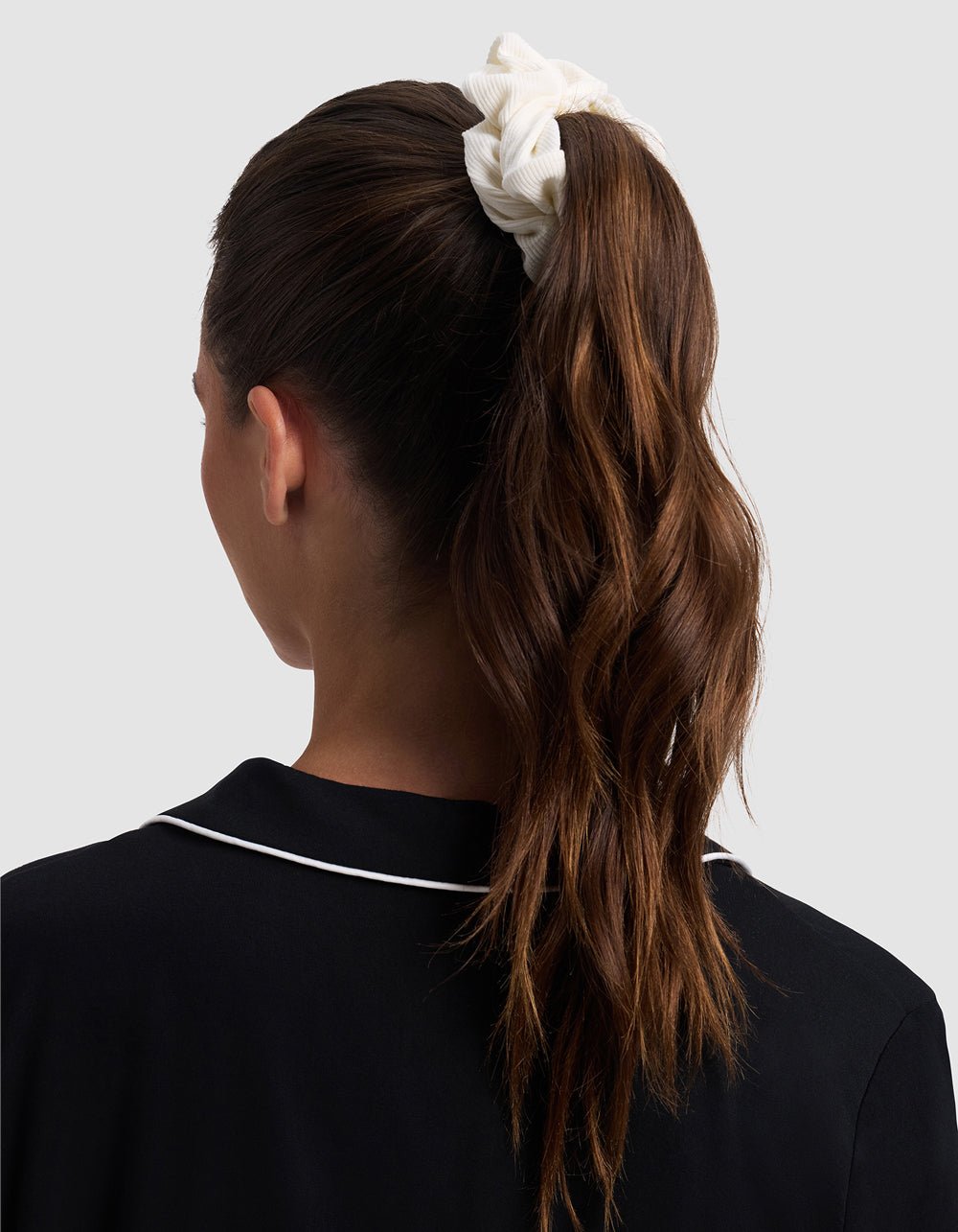 A woman with long, wavy brown hair in a high ponytail, secured with a Cozy Earth Bamboo Rib-Knit Scrunchie, is shown from the back. She wears a black top with white piping along the collar.