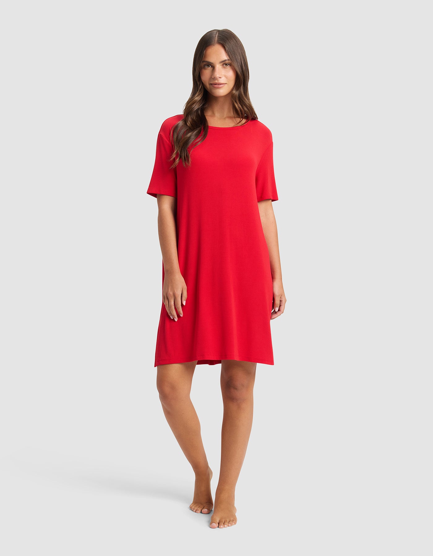 A woman with long brown hair stands barefoot against a light gray background, wearing the Cozy Earth Women’s Bamboo Rib-Knit Boyfriend Sleep Dress in red, featuring short sleeves and a loose fit that falls just above her knees.