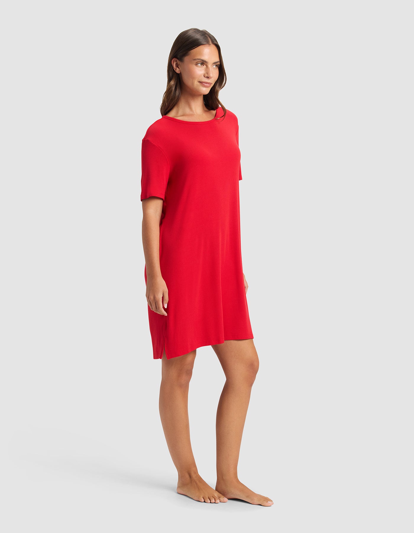 A woman stands barefoot against a light gray background, wearing the Cozy Earth Women’s Bamboo Rib-Knit Boyfriend Sleep Dress in red. She smiles softly and looks to the side.