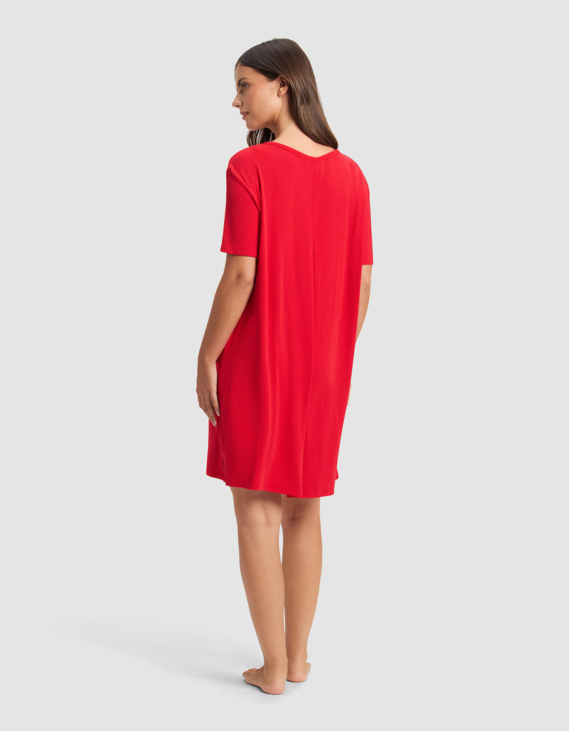 A woman stands barefoot on a plain background, facing away and looking to the side, wearing the Cozy Earth Women’s Bamboo Rib-Knit Boyfriend Sleep Dress—a loose, short-sleeved, knee-length red dress.