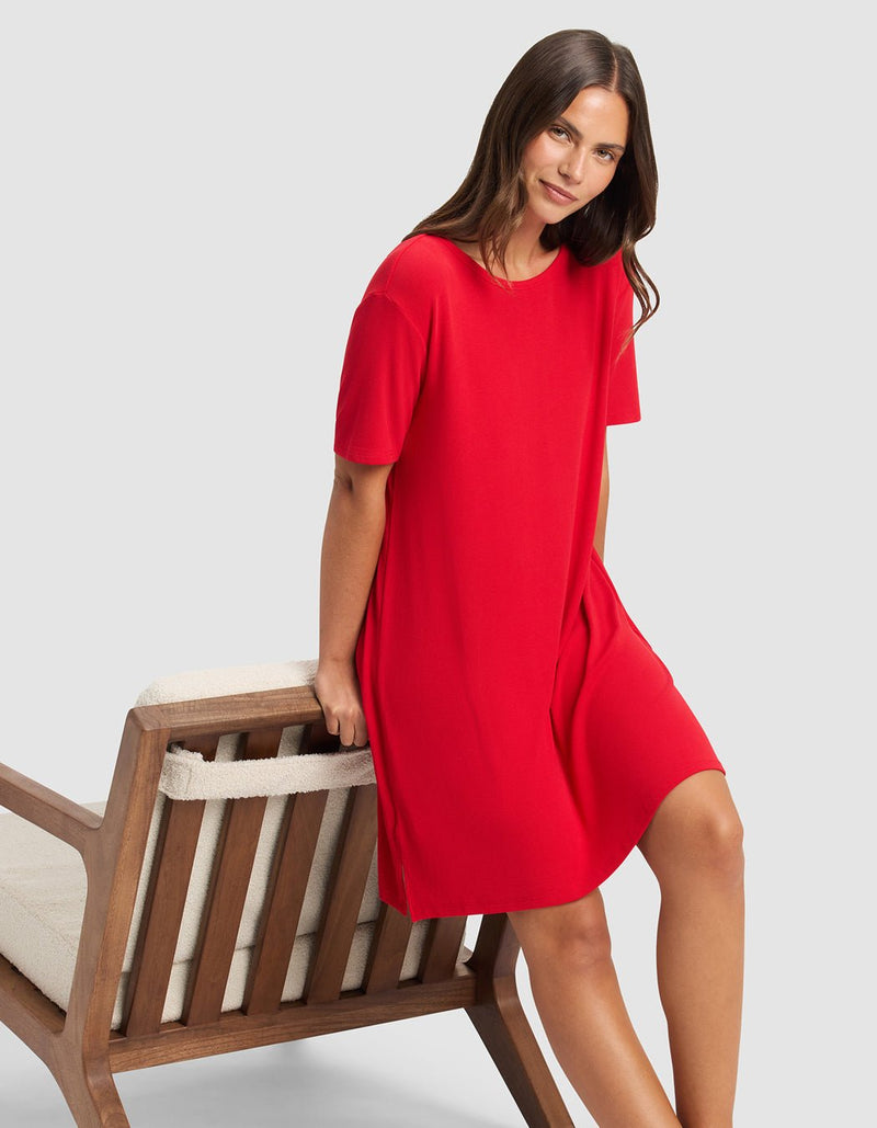 A woman with long brown hair sits on a wooden chair’s armrest, smiling slightly and wearing the Cozy Earth Women’s Bamboo Rib-Knit Boyfriend Sleep Dress in red. The background is plain light gray.
