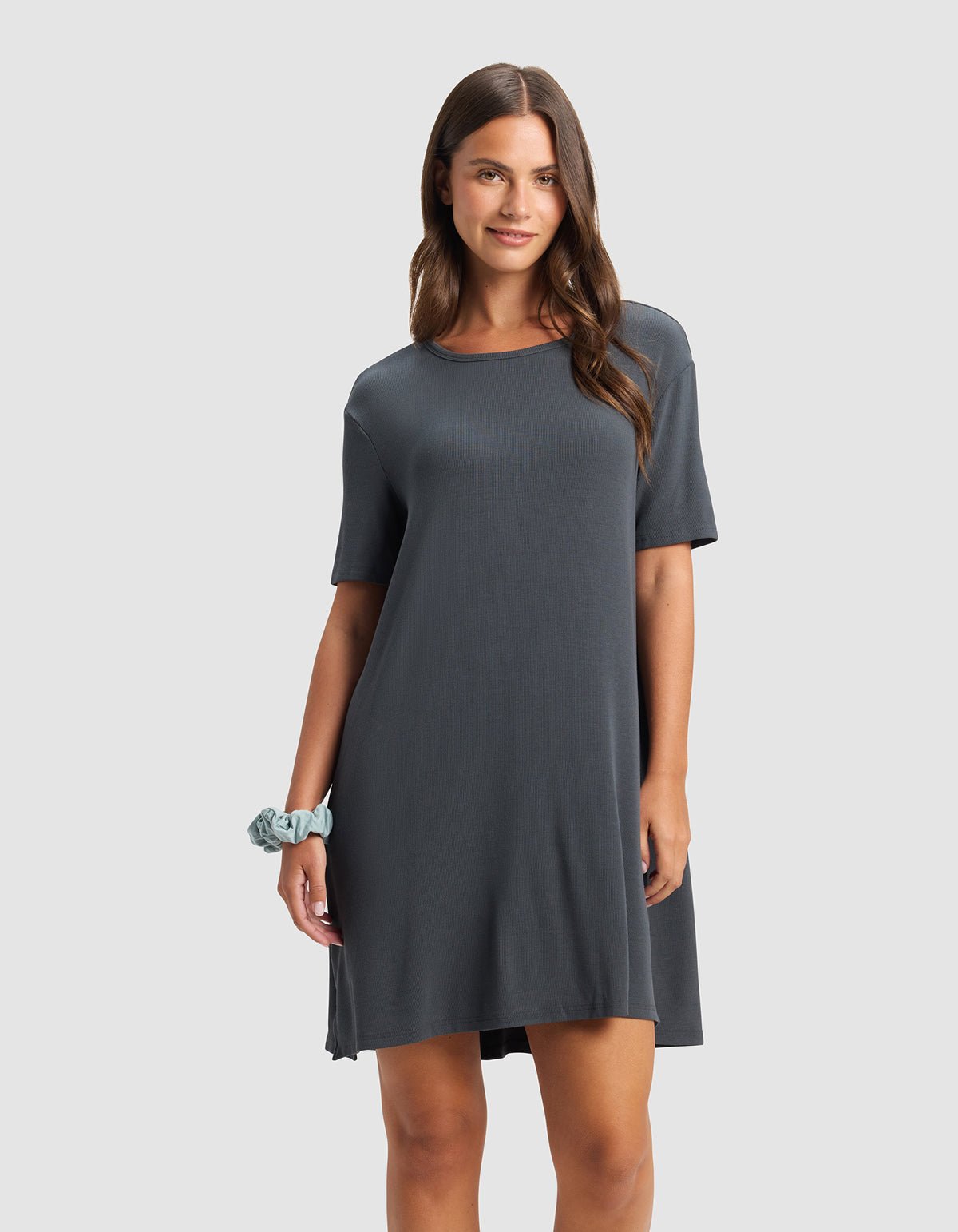 A woman with long brown hair wears the Cozy Earth Women’s Bamboo Rib-Knit Boyfriend Sleep Dress in dark gray and a light blue scrunchie on her wrist. She smiles softly against a plain light gray background.