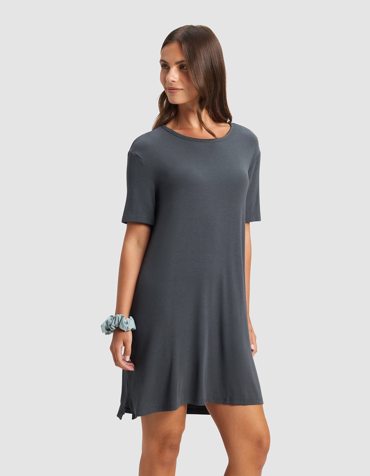 A woman wears the Cozy Earth Women’s Bamboo Rib-Knit Boyfriend Sleep Dress in dark gray, her long brown hair down and a light blue scrunchie on her left wrist, standing against a plain light gray background.