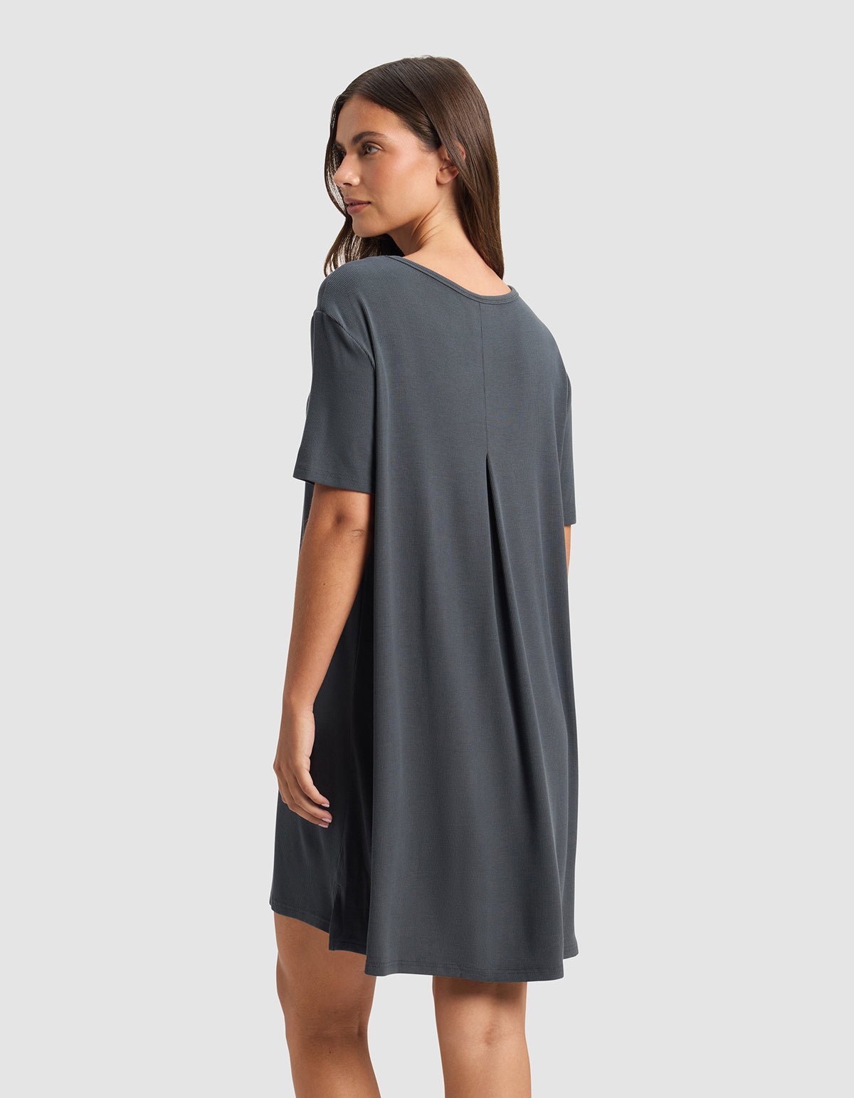 A woman with straight brown hair models the Cozy Earth Women’s Bamboo Rib-Knit Boyfriend Sleep Dress in charcoal gray, viewed from the back. The loose-fitting, short-sleeve dress features a center pleat and falls above the knees against a plain light gray background.