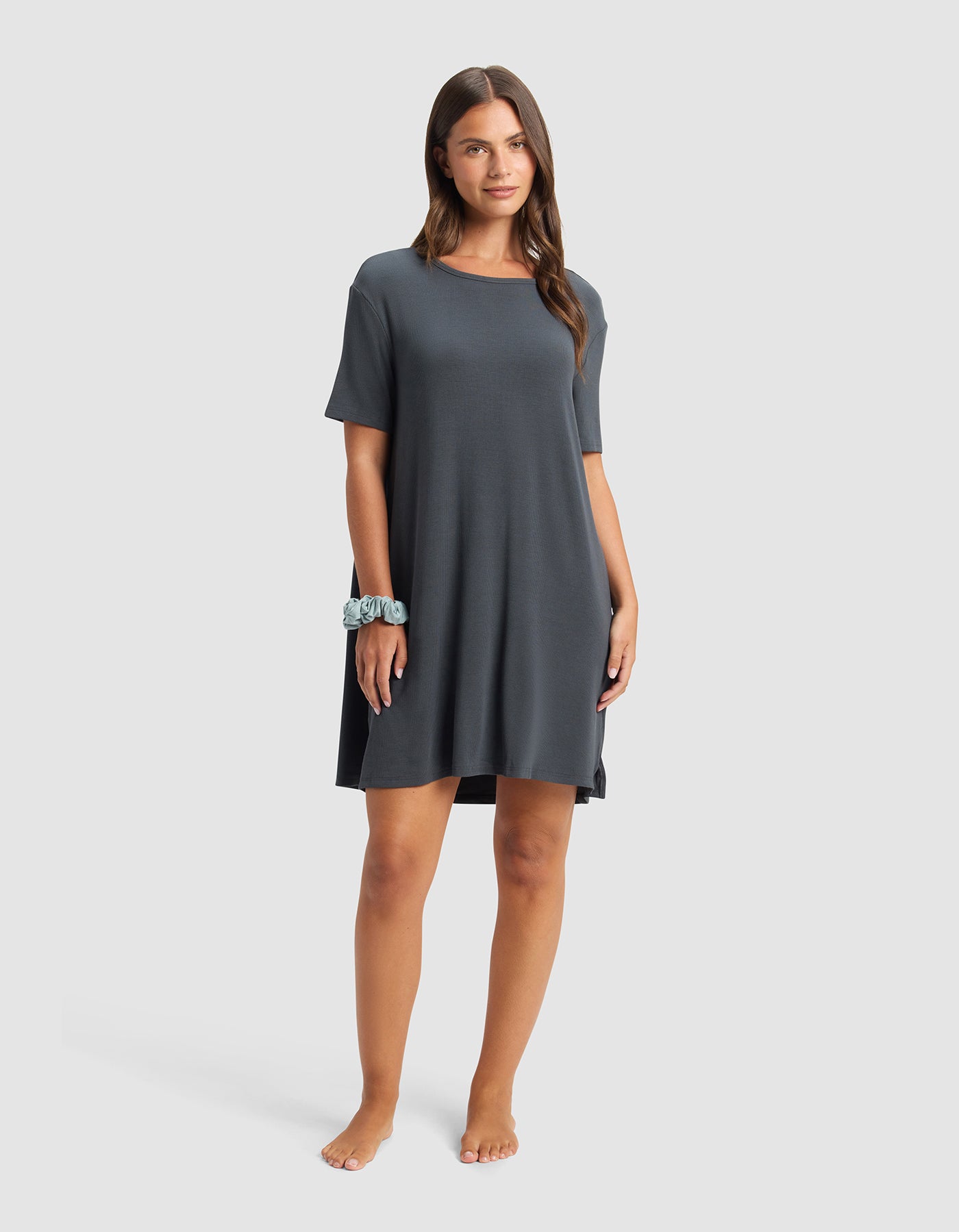A woman with long brown hair stands barefoot against a plain background, wearing the Cozy Earth Women’s Bamboo Rib-Knit Boyfriend Sleep Dress in dark gray, short-sleeved and loose-fitting, with a blue scrunchie on her wrist.