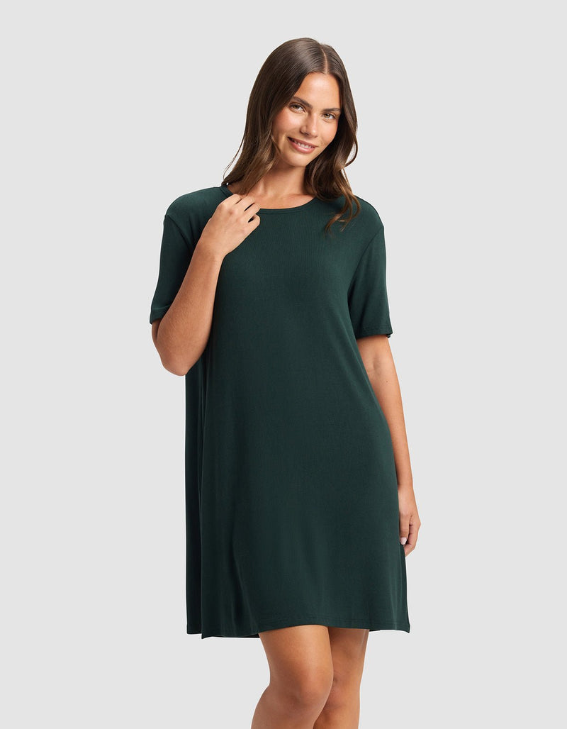 A woman with long brown hair smiles and poses in the Cozy Earth Women’s Bamboo Rib-Knit Boyfriend Sleep Dress, a dark green, short-sleeve, knee-length style, against a plain light gray background.
