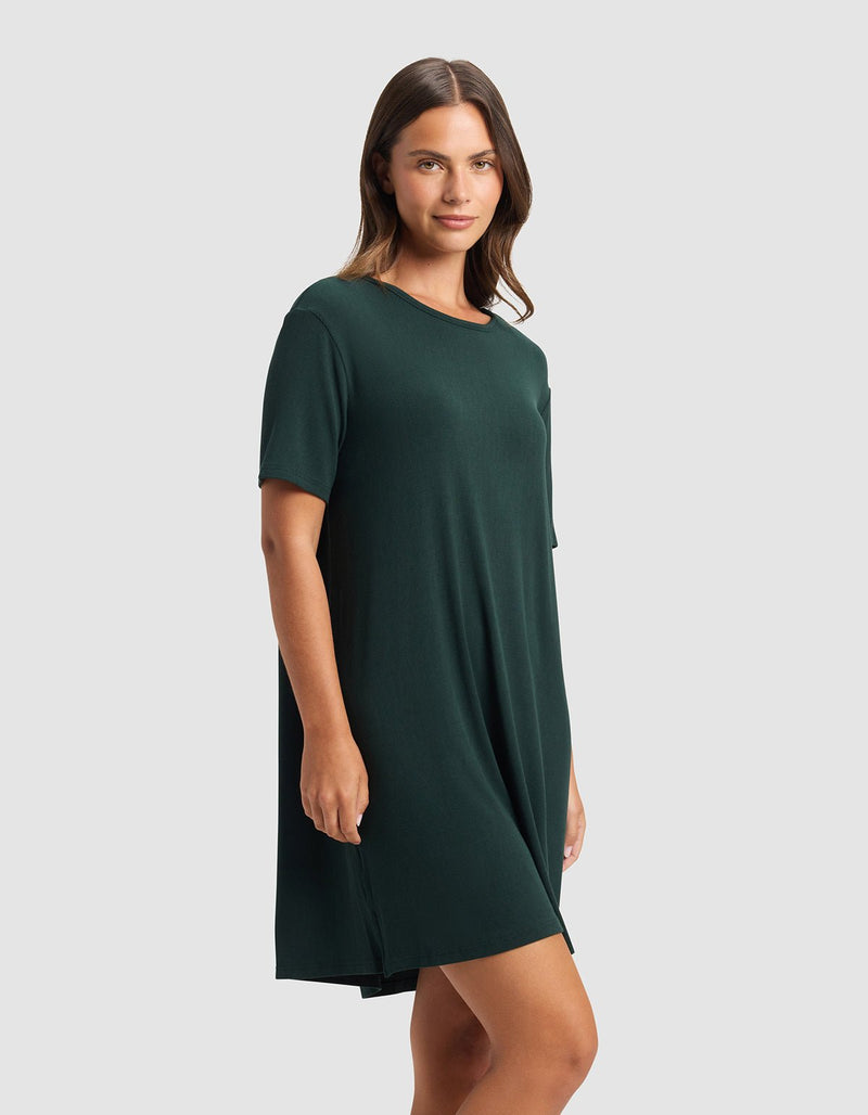 A woman with straight brown hair wears the Cozy Earth Women’s Bamboo Rib-Knit Boyfriend Sleep Dress in dark green, standing against a plain light gray background and looking at the camera with a slight smile.