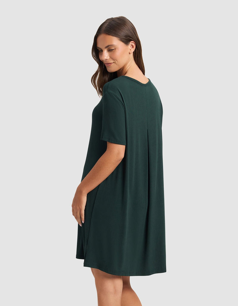 A woman with long brown hair wears the Cozy Earth Women’s Bamboo Rib-Knit Boyfriend Sleep Dress, a loose-fitting, short-sleeved dark green dress, standing sideways and smiling slightly against a plain light gray background.