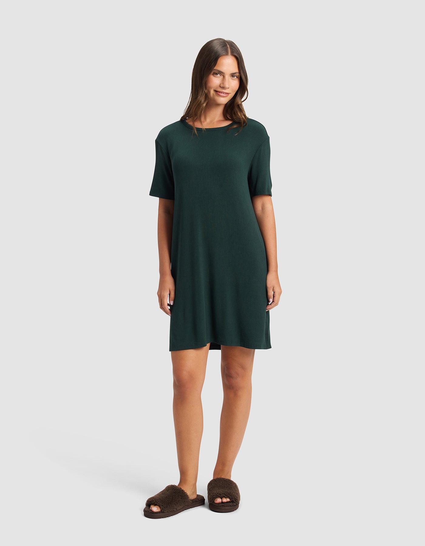 A woman smiles, wearing the Cozy Earth Women's Bamboo Rib-Knit Boyfriend Sleep Dress and brown fuzzy slippers, standing against a plain light gray background.|Color:Spruce