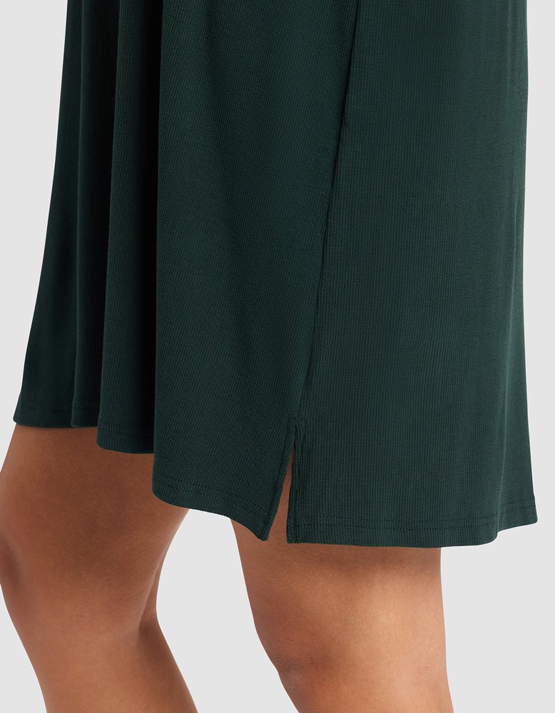 A close-up of a person wearing the Cozy Earth Women’s Bamboo Rib-Knit Boyfriend Sleep Dress in dark green, featuring a small side slit that reveals part of their upper leg against a plain, light background.