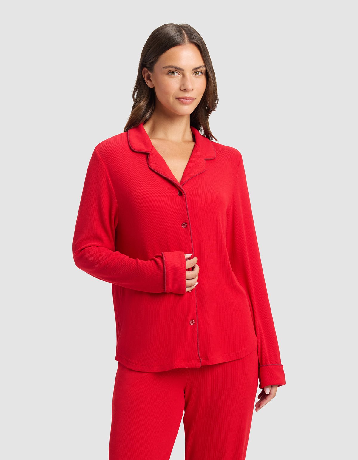 A woman with long brown hair stands against a light gray background, wearing the Cozy Earth Women's Bamboo Rib-Knit Classic Long Sleeve Pajama Top in red and matching pants. She smiles gently with one hand on her abdomen.