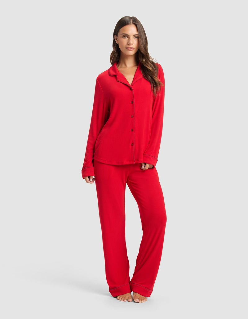 A woman stands barefoot against a light gray background, wearing Cozy Earth's Women's Bamboo Rib-Knit Classic Long Sleeve Pajama Top in red with matching loose-fitting pants.