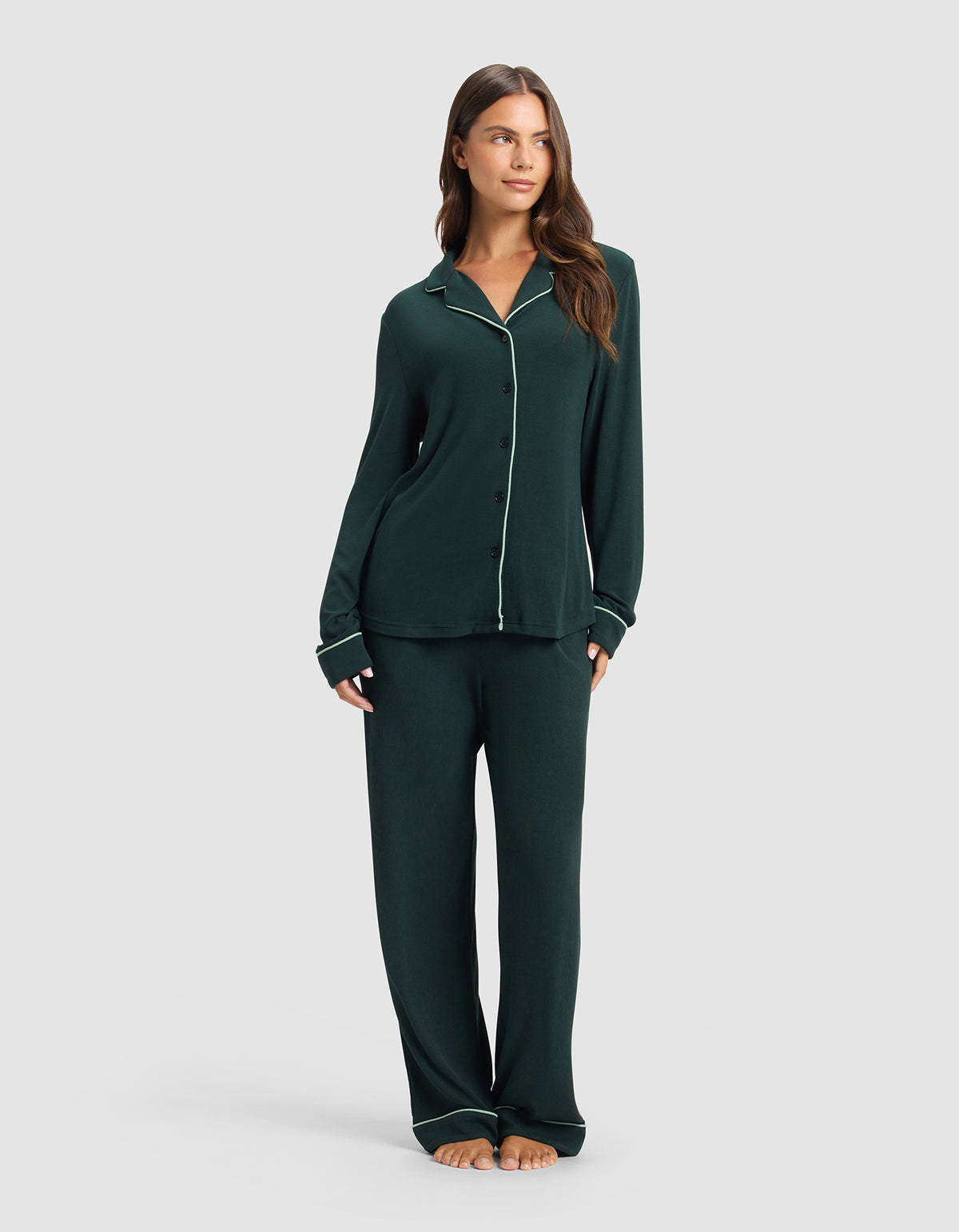 A woman stands barefoot against a light gray background, wearing Cozy Earth's Women's Bamboo Rib-Knit Classic Long Sleeve Pajama Top in dark green with white piping, paired with matching loose pants.|Color:Spruce