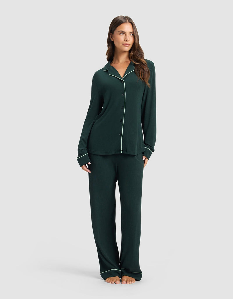 A woman stands barefoot against a light gray background, wearing Cozy Earth's Women's Bamboo Rib-Knit Classic Long Sleeve Pajama Top in dark green with white piping, paired with matching loose pants.