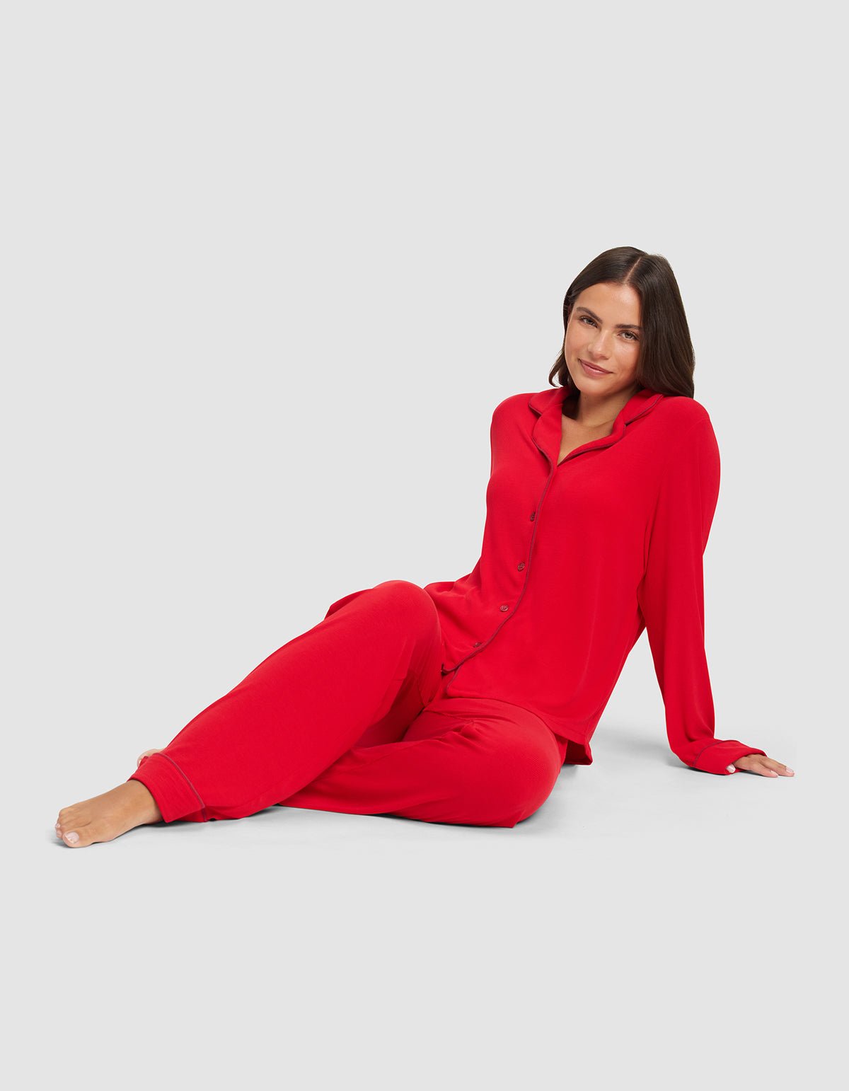 A dark-haired woman sits barefoot on the floor against a light gray background, wearing Cozy Earth's Women's Bamboo Rib-Knit Classic Pajama Pants in bright red with a relaxed, calm expression and a matching button-up shirt.