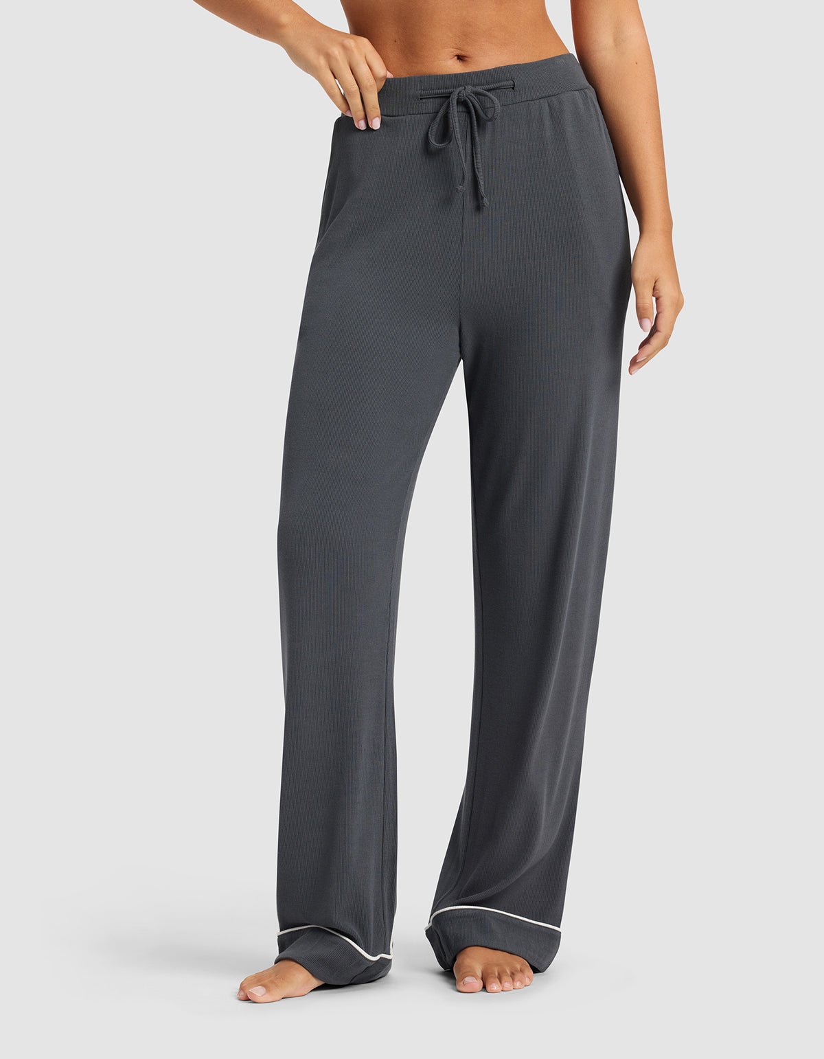 A woman in Cozy Earth Women's Bamboo Rib-Knit Classic Pajama Pant in dark gray, featuring a drawstring waist and white piping at the cuffs, stands barefoot against a plain background.|Color:Graphite