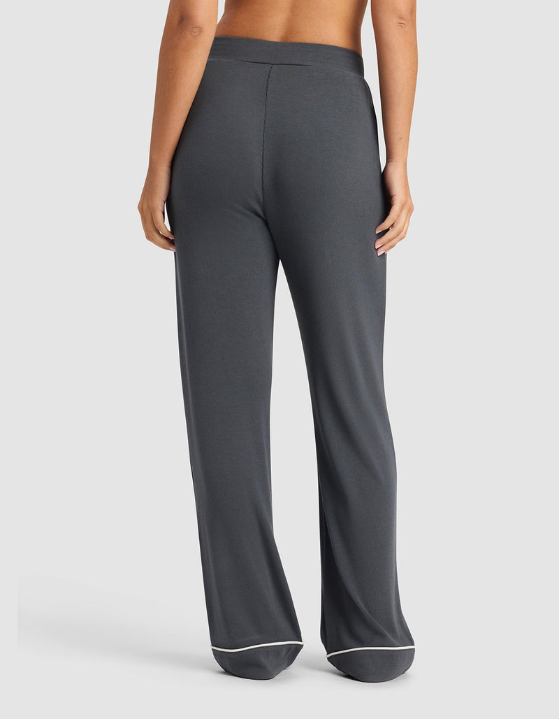 A woman is shown from the back wearing Cozy Earth's Women's Bamboo Rib-Knit Classic Pajama Pant in dark gray with white trim, standing barefoot against a plain light background.